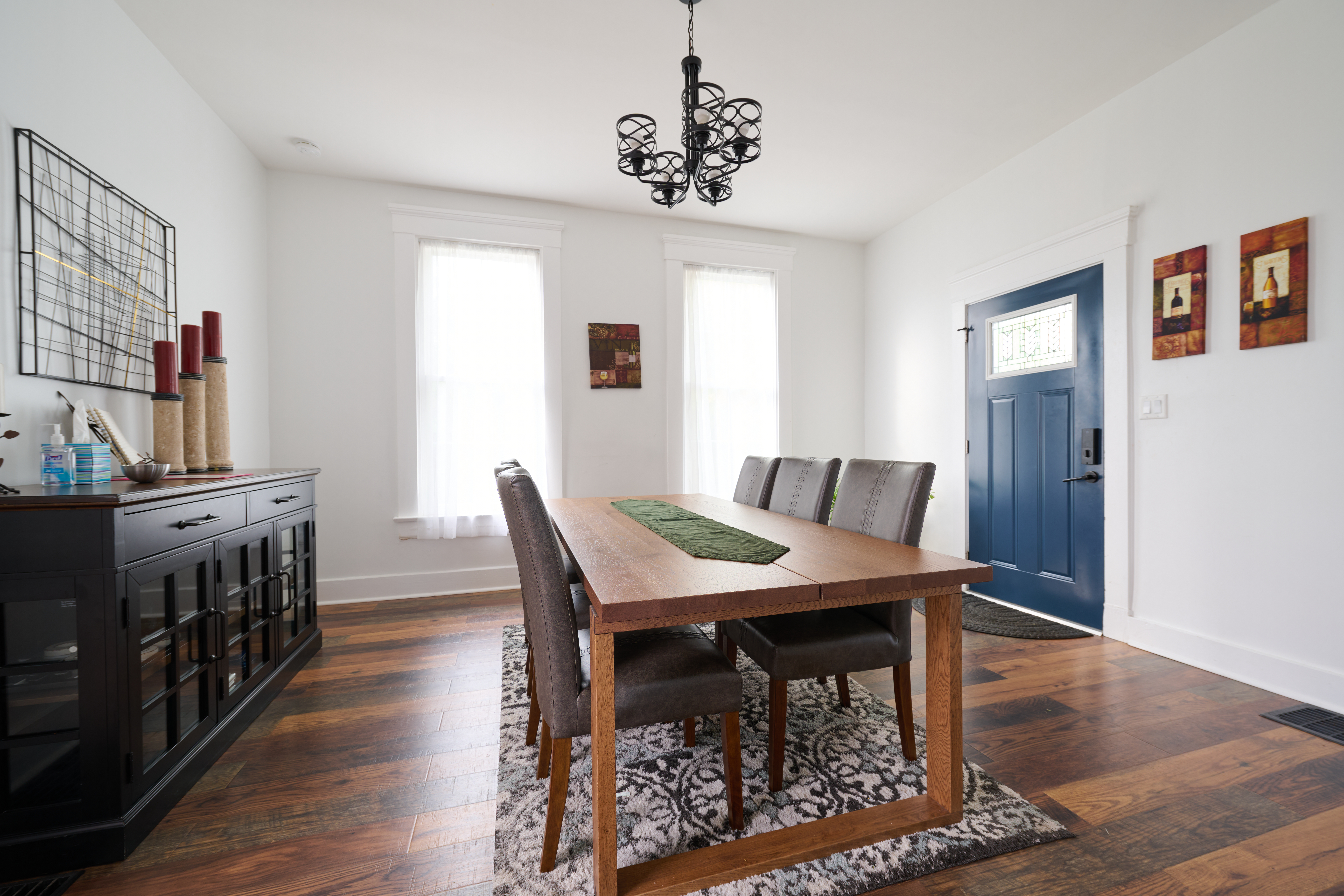 A bright dining room with a wooden table, six gray upholstered chairs, a blue door, and decorative wall art under a black chandelier.