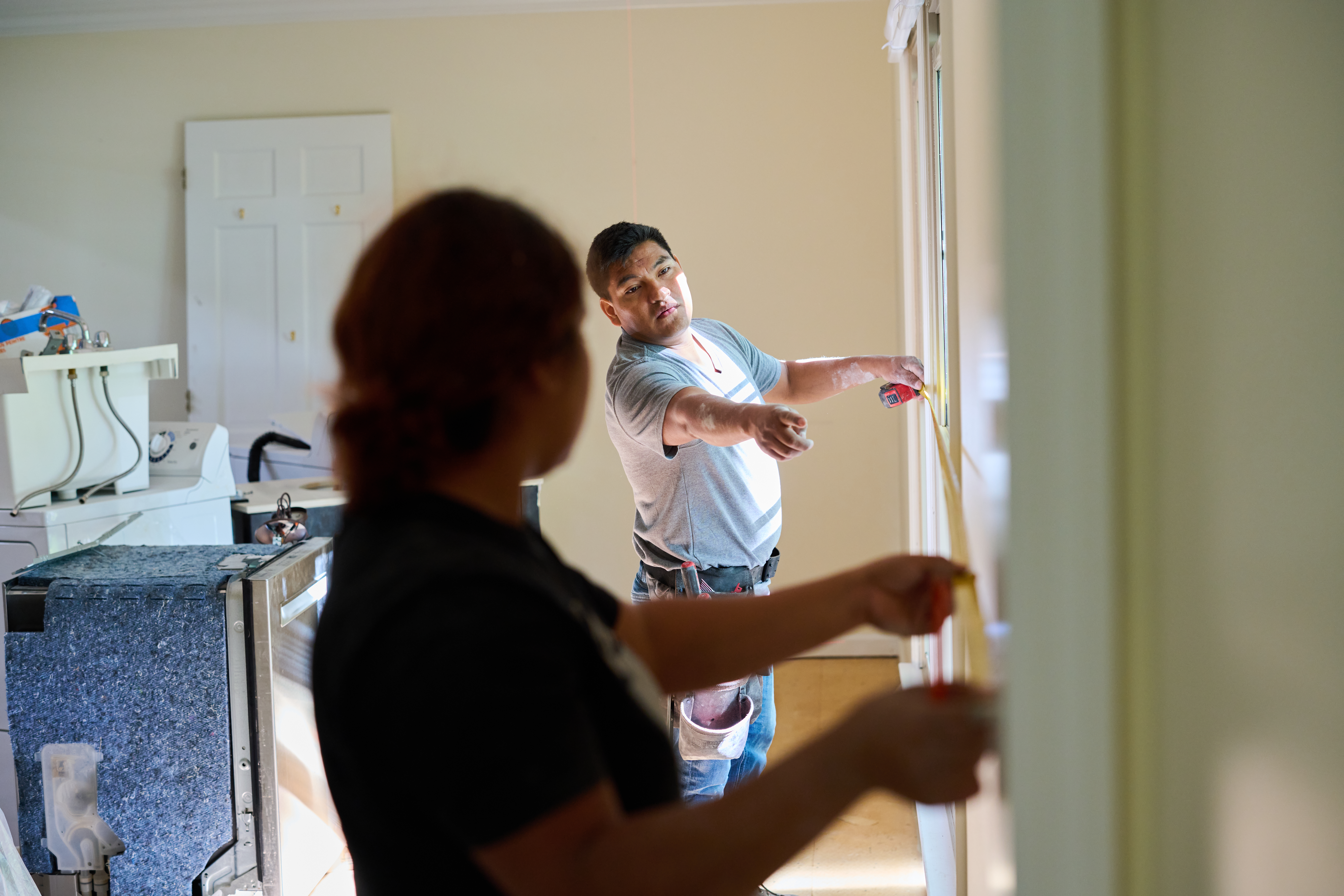 Two workers measuring a wall with a tape measure in a bright room, discussing placement for a renovation project.